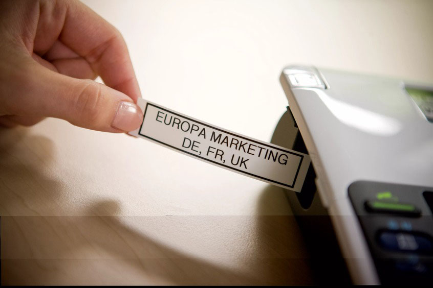 Mano sosteniendo una etiqueta blanca ancha con el texto “EUROPA MARKETING DE, FR, UK” junto a una rotuladora Brother P-touch.