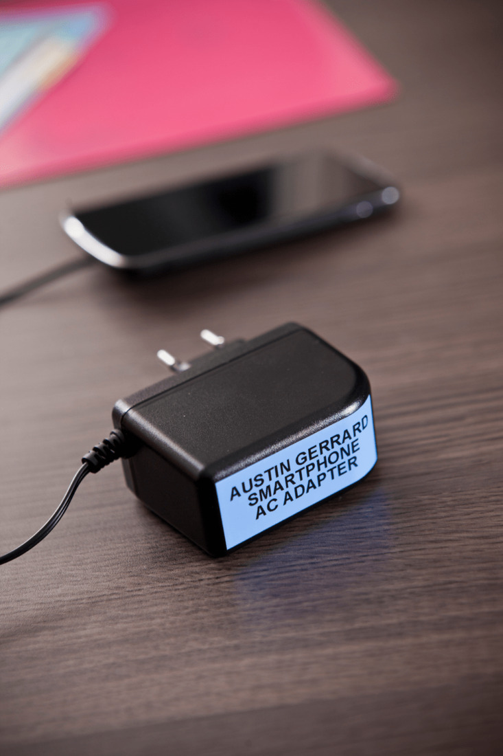 Adaptador de corriente negro para smartphone identificado con una etiqueta azul que dice “Austin Gerard Smartphone AC Adapter”.