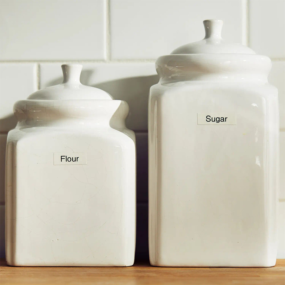 Recipientes de cocina blancos identificados con etiquetas Brother TZe-231 para harina y azúcar.