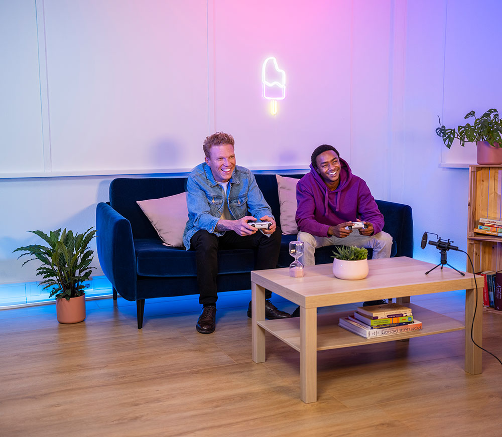 Dos personas sentadas en un sofá azul jugando videojuegos con controles inalámbricos en una sala de estar iluminada con luces de neón rosas y azules, con un Micrófono USB para streaming Trust GXT 241 Velica, una planta y libros sobre una mesa de centro frente a ellos.