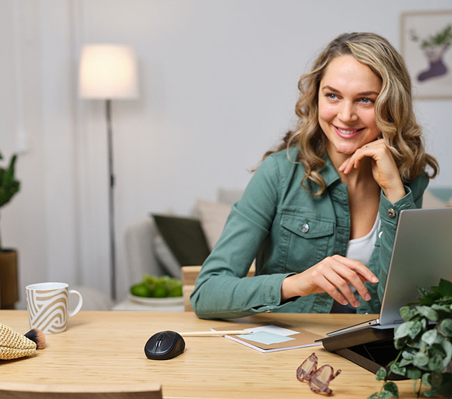 Una persona sentada frente a una computadora portátil en un escritorio con un Mouse inalámbrico compacto Bluetooth® Trust Primo, una taza, anteojos y una planta decorativa alrededor, en un ambiente interior iluminado.