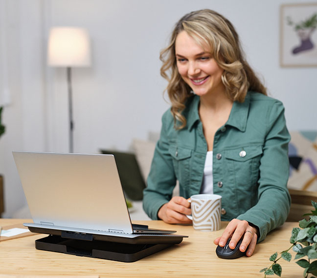 Persona con camisa verde sentada frente a una laptop sobre un escritorio, utilizando un mouse inalámbrico compacto Bluetooth® Trust Primo y sosteniendo una taza. La escena incluye una lámpara y plantas decorativas, en un ambiente de oficina o casa con estilo moderno.