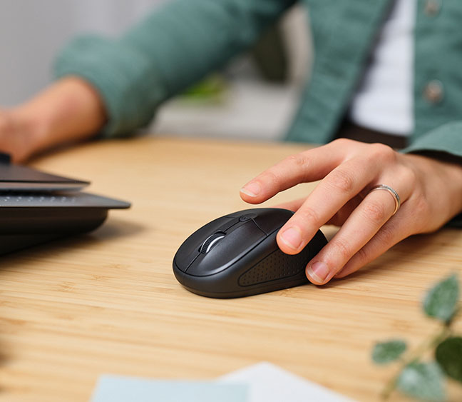 Imagen de un mouse inalámbrico compacto Bluetooth® Trust Primo sobre una mesa de madera, junto a una mano de una persona con camisa verde, un teclado parcialmente visible y hojas sueltas.