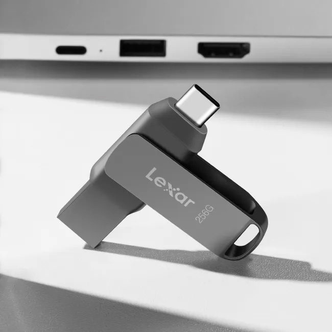 Pendrive Lexar JumpDrive Dual Drive D400 de 256 GB color gris oscuro con un conector USB-C visible, apoyado sobre una superficie blanca frente a una laptop