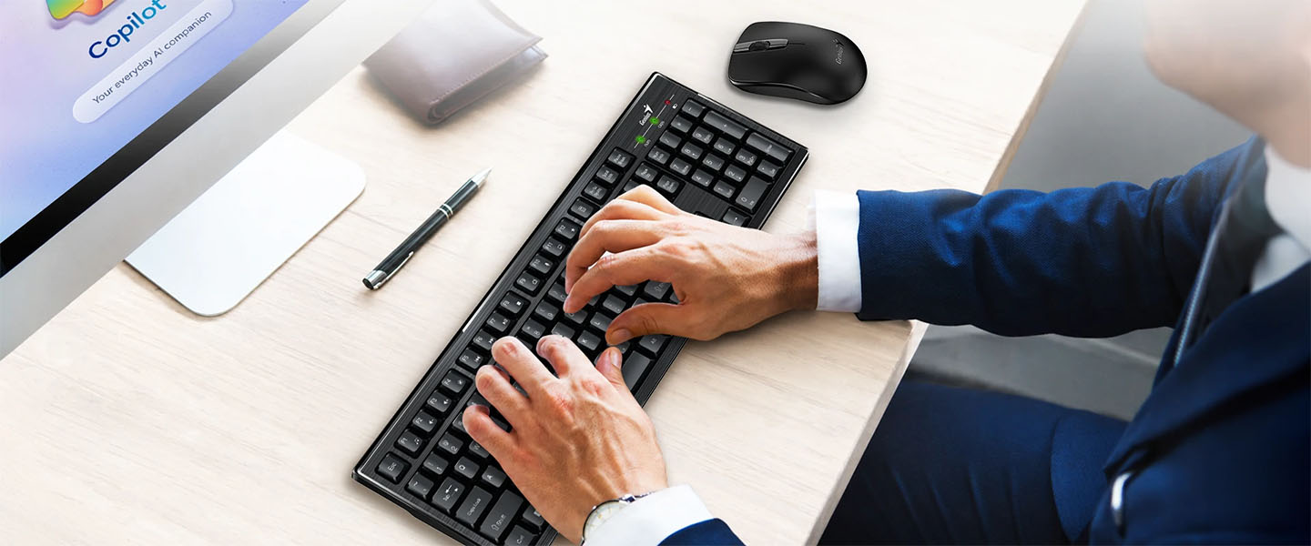 Persona usando el teclado Genius KM-8101 frente a una computadora con Copilot en pantalla y mouse inalámbrico sobre el escritorio