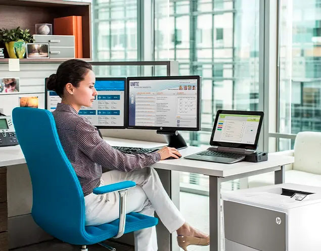 Mujer usando una impresora HP Color LaserJet Pro 4203dw en un escritorio con monitores dobles y silla azul