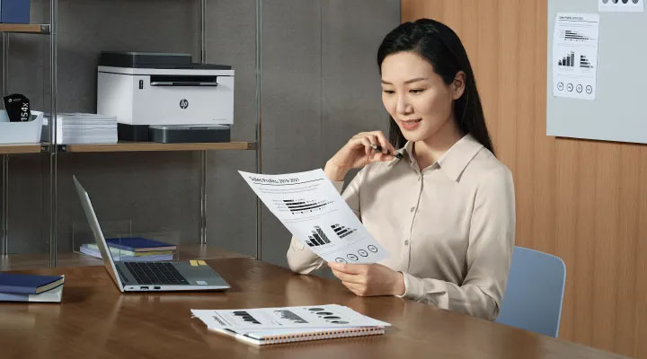 Impresora multifunción HP LaserJet Tank blanca en estante de oficina mientras una mujer revisa gráficos impresos.