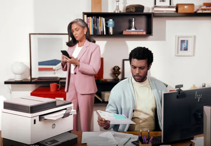 Impresora multifunción HP LaserJet Tank MFP 2602sdw blanca en ambiente de oficina moderna, usada por un hombre mientras una mujer usa su móvil.