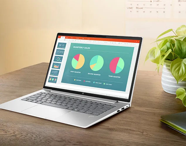 Laptop HP ProBook 445 G11 de 14 pulgadas plateada abierta sobre un escritorio de madera. Muestra en pantalla un gráfico de Ventas Trimestrales con tres diagramas circulares. Se ve parcialmente una planta de interior y un pizarrón en el fondo.