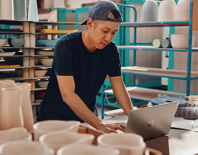 Hombre joven usando una laptop HP ProBook 445 G11 en un taller de cerámica. Lleva puesta una gorra y una remera oscura mientras trabaja concentrado. Se ven estanterías con piezas de cerámica sin terminar a su alrededor.