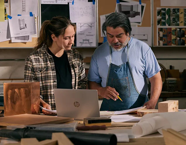 Dos personas trabajando en un taller con una laptop HP ProBook 445 G11 abierta sobre el escritorio. Un hombre con delantal azul y barba canosa señala planos con un lápiz mientras una mujer con camisa a cuadros mira la pantalla. Hay herramientas y materiales de carpintería y diseño en primer plano.