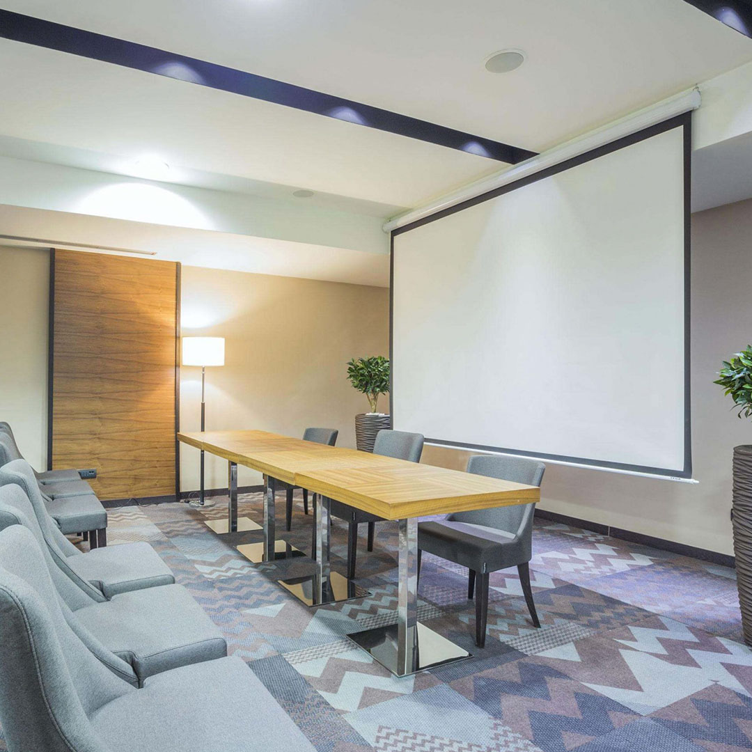 Sala de conferencias moderna con una pantalla para proyección de imagen con bloqueo automático Intelaid IT-SPS100. mesa de madera con sillas a juego y sillones grises en primer plano. El espacio cuenta con iluminación empotrada y una lámpara de pie, sobre un suelo alfombrado con patrón geométrico.