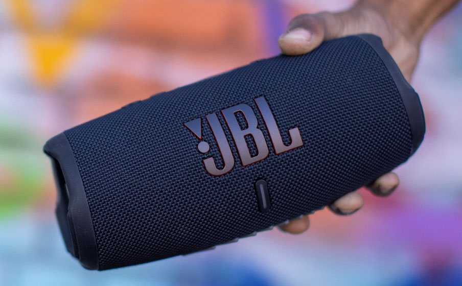 Primer plano de un parlante Bluetooth JBL Charge 5 negro sostenido por una mano, con un fondo de arte abstracto borroso.