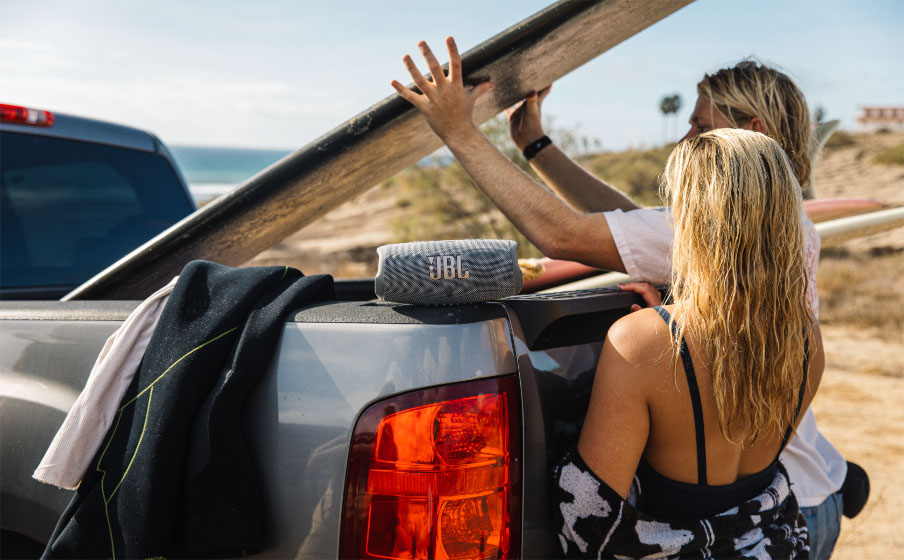 Parlante Bluetooth JBL Charge 5 gris sobre la caja de una camioneta mientras una pareja rubia carga tablas de surf en la playa.