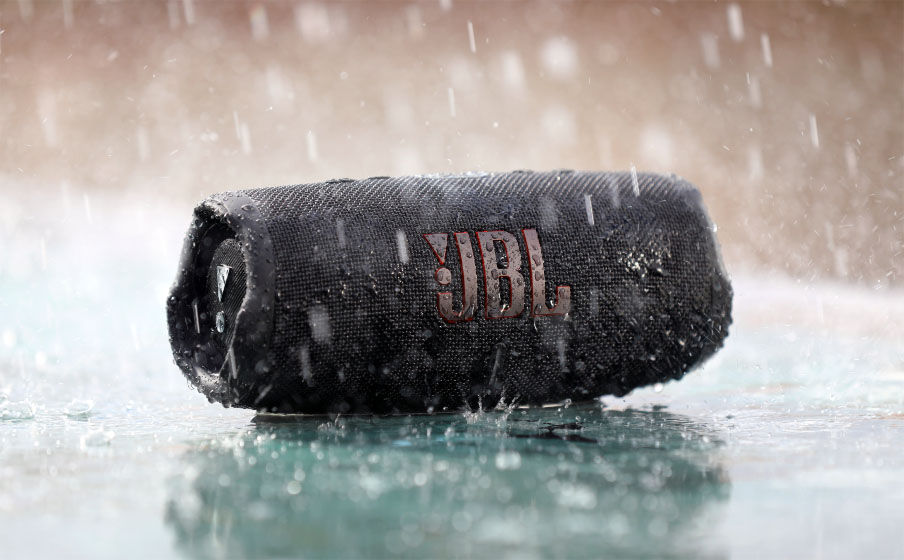 Parlante Bluetooth JBL Charge 5 negro resistiendo una lluvia fuerte, sobre una superficie mojada.
