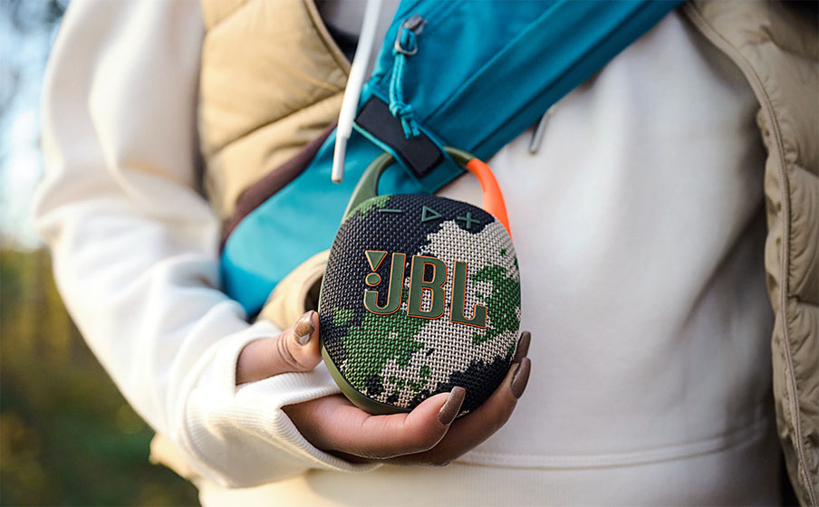 Persona sosteniendo el parlante Bluetooth JBL Clip 5 con patrón de camuflaje en sus manos, con una correa de bolso turquesa cruzada al fondo.