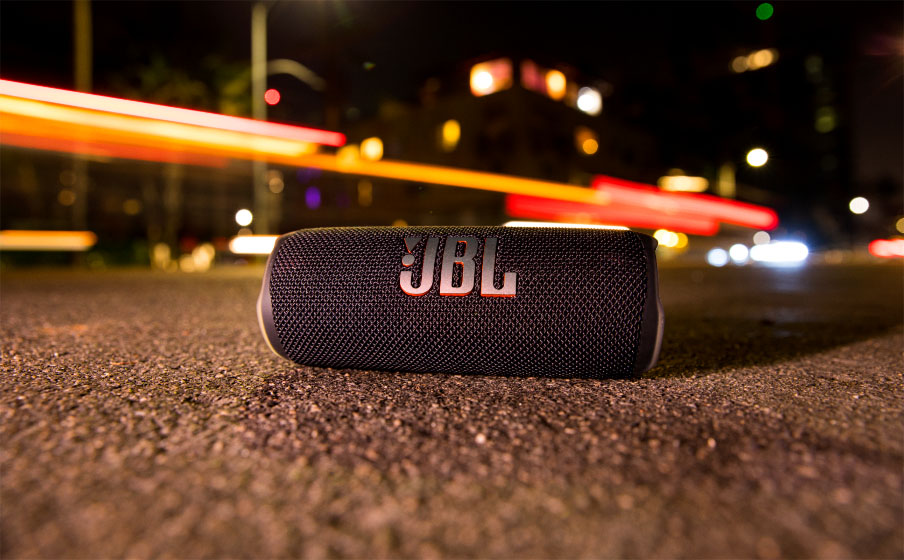Parlante Bluetooth JBL Flip 6 negro sobre asfalto de noche, con luces de coches borrosas y brillantes de fondo.