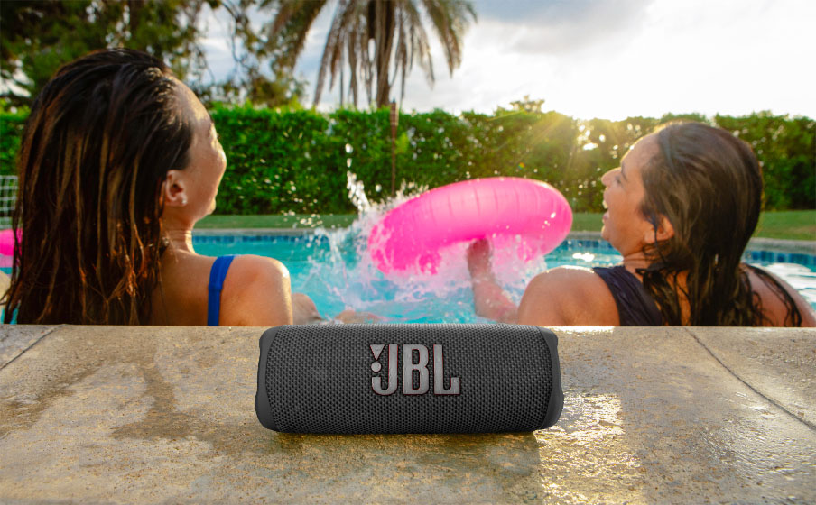 Parlante Bluetooth JBL Flip 6 gris sobre el borde de una pileta con dos mujeres salpicando agua y divirtiéndose atrás.