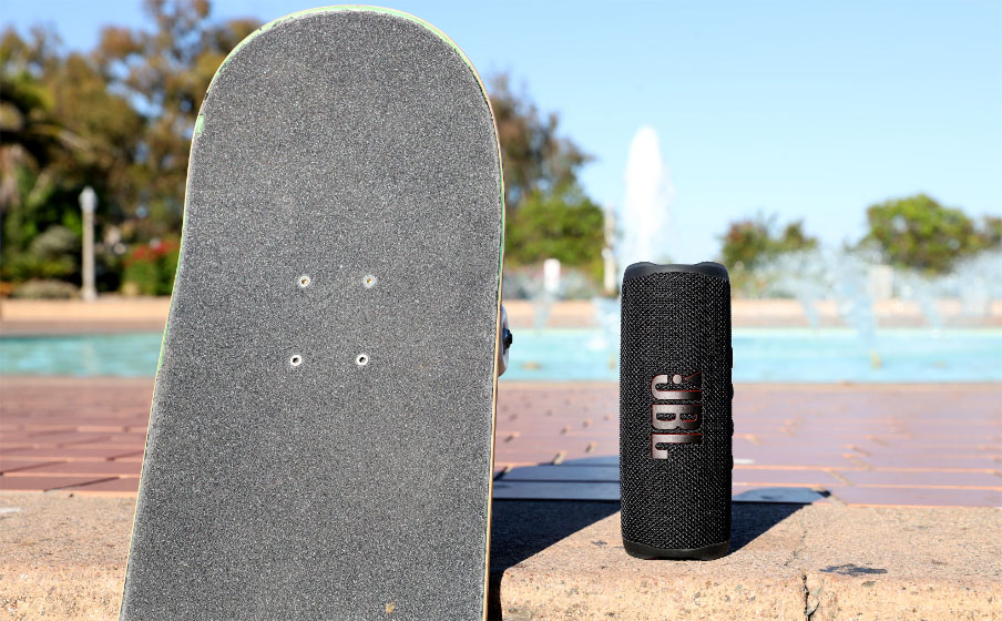Parlante Bluetooth JBL Flip 6 negro al lado de una patineta de pie, en un parque con una fuente de agua de fondo.