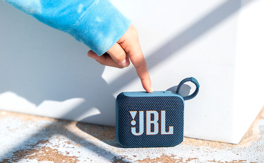 Parlante Bluetooth JBL Go 4 azul sobre arena, una mano presionando el botón superior.