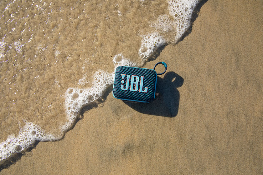 Parlante Bluetooth JBL Go 4 azul sobre arena mojada en la playa, con una ola espumosa llegando hasta él.
