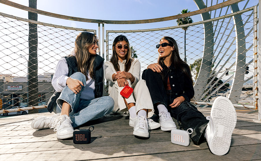 Tres chicas jóvenes sentadas y riendo al aire libre, con los parlantes Bluetooth JBL Go 4 en negro, rojo y blanco frente a ellas.