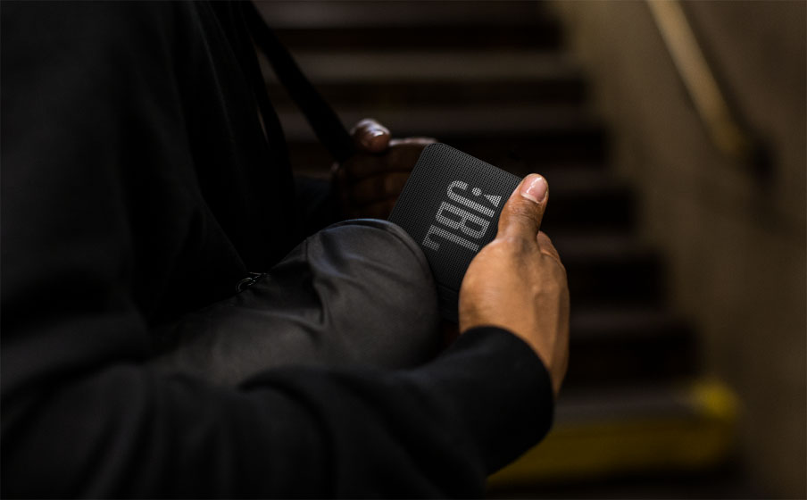 Manos de una persona poniendo el parlante Bluetooth JBL Go Essential negro en un bolsillo, con unas escaleras oscuras de fondo.