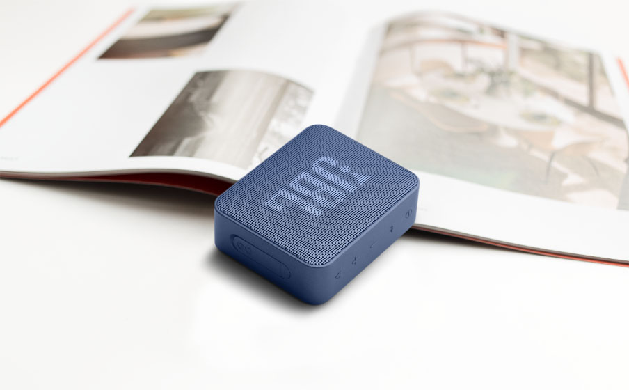 Parlante Bluetooth JBL Go Essential azul sobre una mesa blanca con una revista abierta en el fondo.