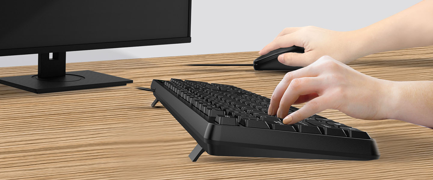 Una persona utilizando un Combo de Teclado y Mouse Genius KM-170 sobre una mesa de madera frente a un monitor de computadora