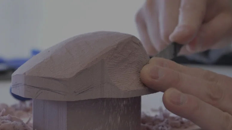Manos tallando una figura de madera con forma de mouse utilizando una herramienta de corte.