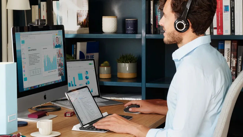 Hombre con auriculares participando en una videollamada desde una oficina en casa, usando una computadora de escritorio, una notebook y una tablet al mismo tiempo.
