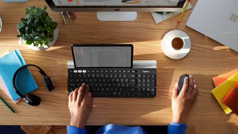 Vista superior de una persona trabajando en un escritorio con un teclado y un Mouse Logitech M720 Triathlon, usando una tablet como pantalla principal entre una taza de café, auriculares y artículos de oficina.