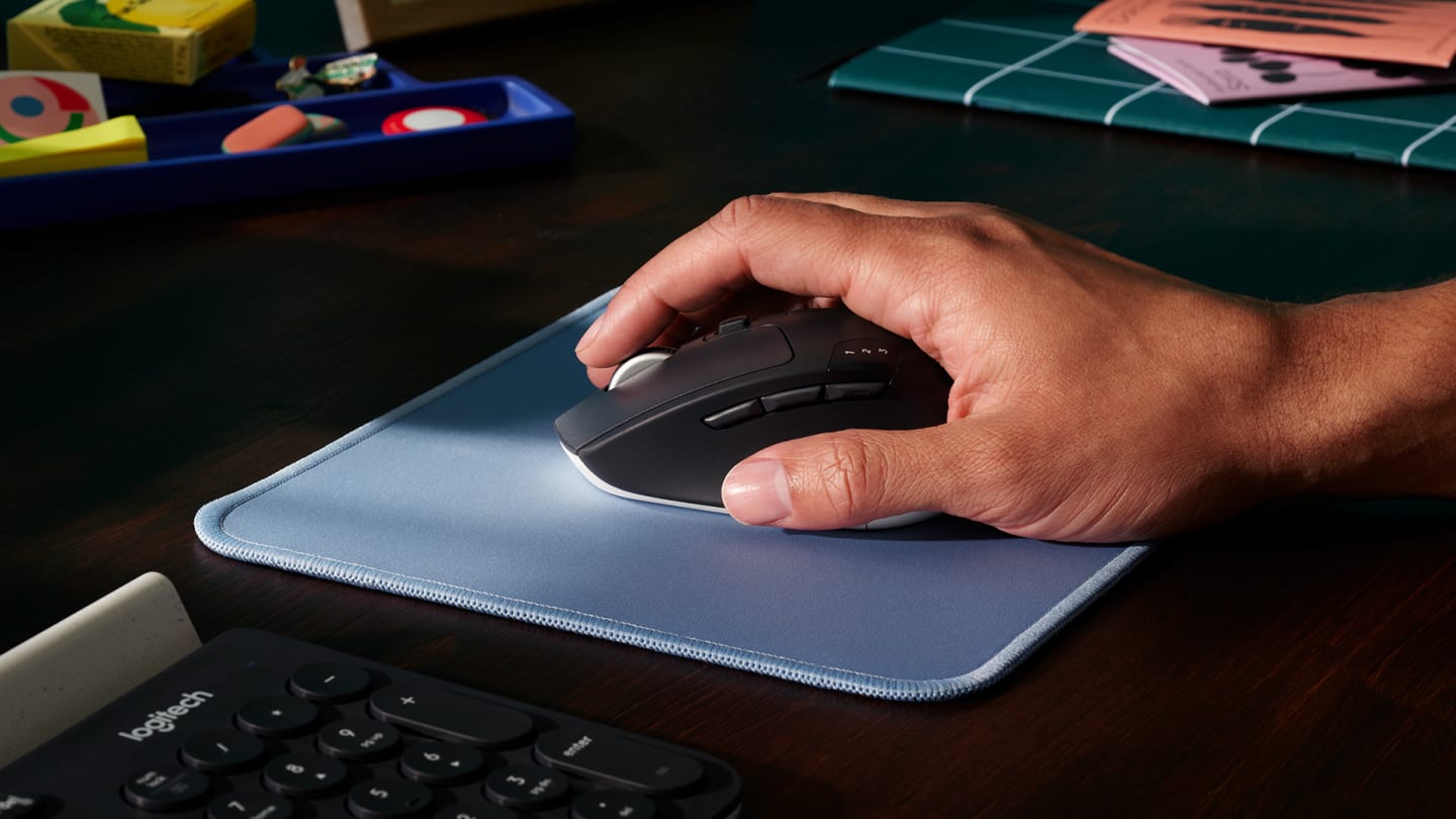 Mano usando un Mouse Logitech M720 Triathlon sobre un mousepad azul, con un teclado y útiles escolares alrededor.