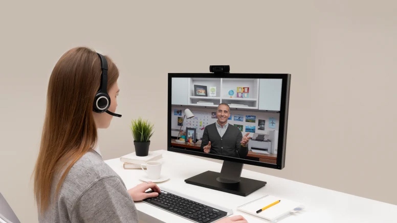 Mujer con auriculares participando en una videollamada desde su escritorio, mirando a un hombre que aparece en la pantalla del monitor.