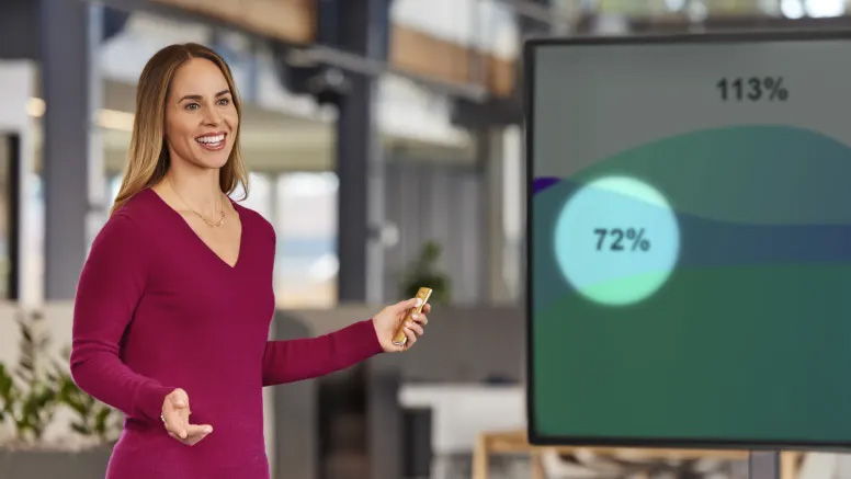 Mujer dando una presentación con un control Remoto Para Presentaciones Logitech Spotlight en la mano, señalando una pantalla que muestra gráficos con porcentajes.