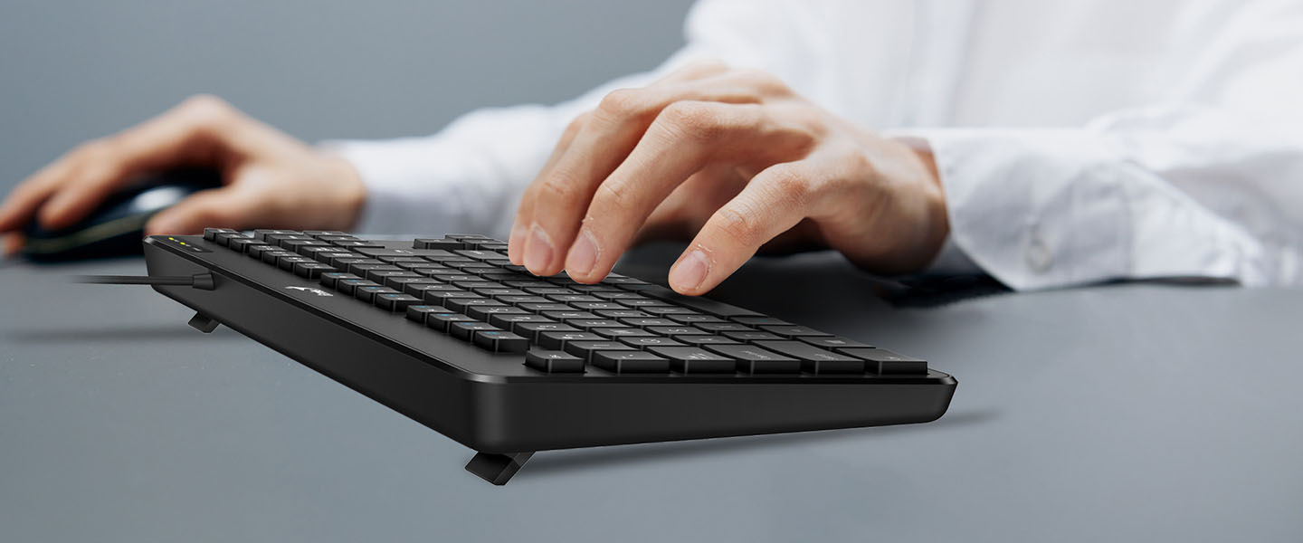Una mano descansa sobre un Teclado Genius LuxeMate 110 USB Negro con fondo gris.