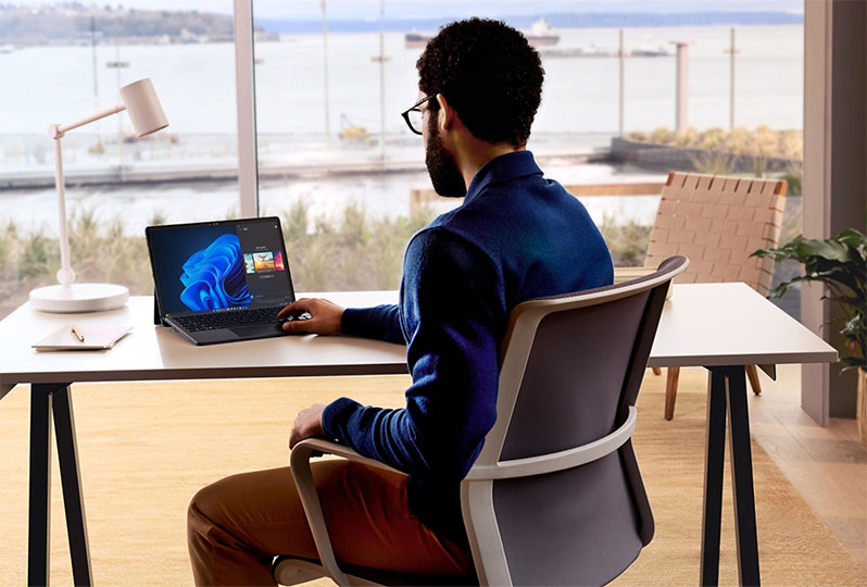 Un hombre está sentado y trabajando en un dispositivo 2 en 1 de Microsoft desde una oficina mirando al agua.