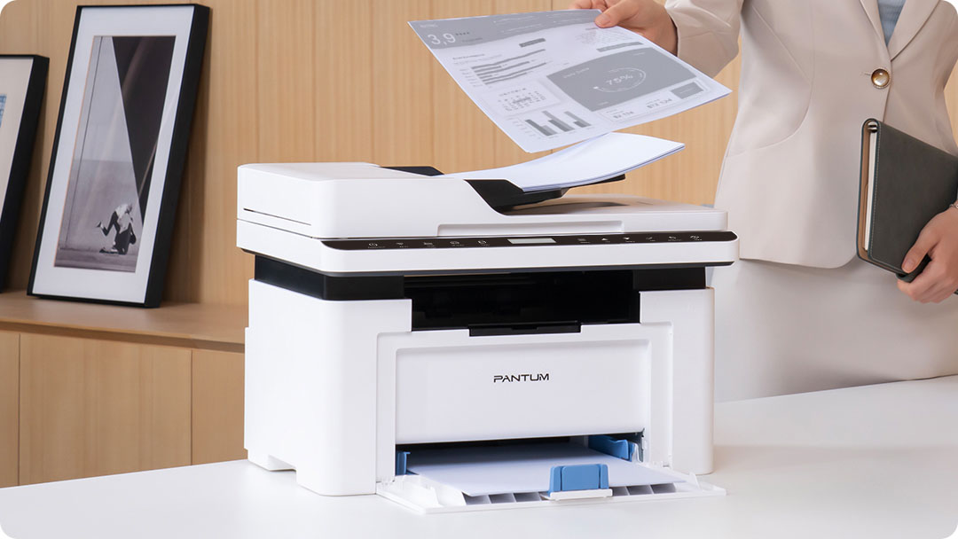 Una persona sostiene hojas de papel sobre una impresora multifunción Pantum BM2300AW blanca y negra en un escritorio blanco, en una oficina con decoración moderna.