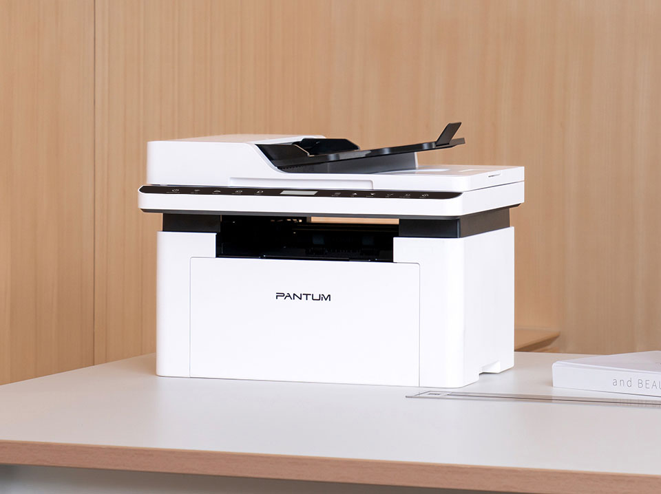 Una impresora multifunción Pantum BM2300AW blanca y negra sobre una mesa clara con un fondo de madera clara.