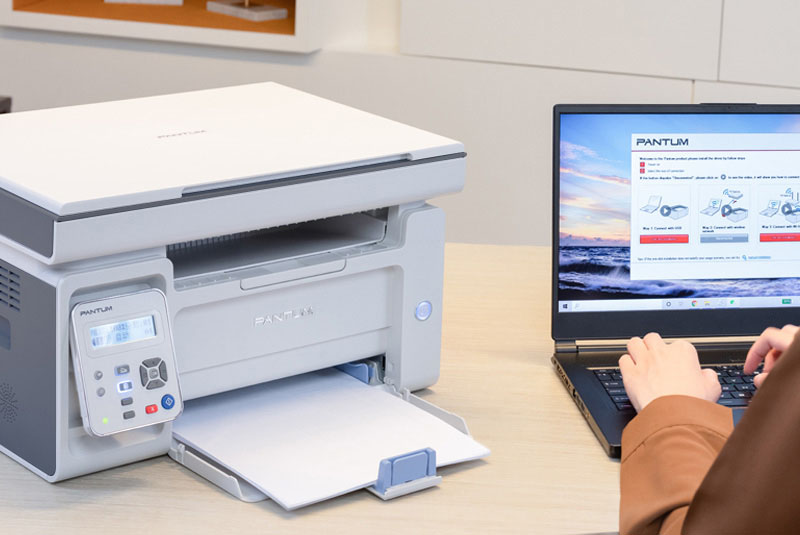 Una impresora multifunción Pantum M6509NW blanca y gris junto a una laptop abierta que muestra la página web de Pantum. Una persona usa la laptop.
