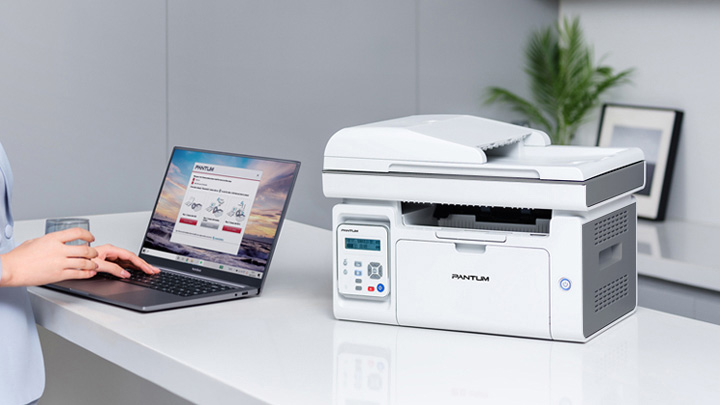 Una impresora multifunción Pantum M6559NW blanca y gris sobre un mostrador blanco, junto a una laptop abierta que muestra la página web de Pantum. Se ven manos usando la laptop.