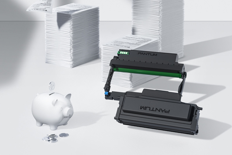 Un cartucho de tóner Pantum negro y verde al lado de una alcancía blanca con monedas, y pilas de hojas de papel en el fondo.