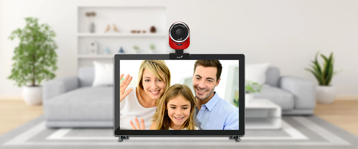 Webcam Genius QCam 6000 instalada encima de una pantalla de TV en una sala de estar, mostrando a una familia sonriente (padre, madre e hija) saludando en una videollamada