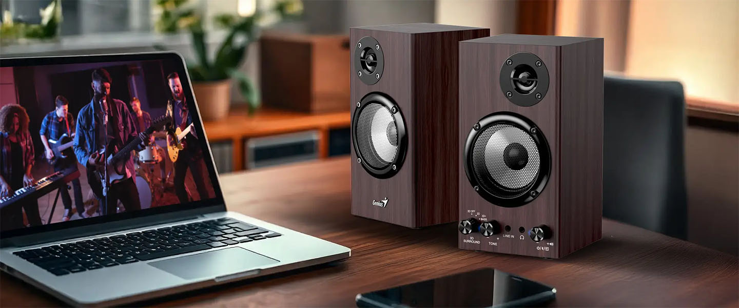 Par de parlantes Genius SP-HF520BT de madera oscura junto a una notebook que reproduce un video musical, todo sobre un escritorio en un ambiente cálido y moderno