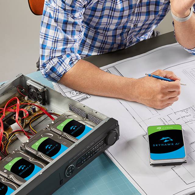 Persona trabajando en un escritorio con planos arquitectónicos, sosteniendo un lápiz azul. Hay un disco duro externo Seagate SkyHawk AI y una computadora con componentes electrónicos visibles.