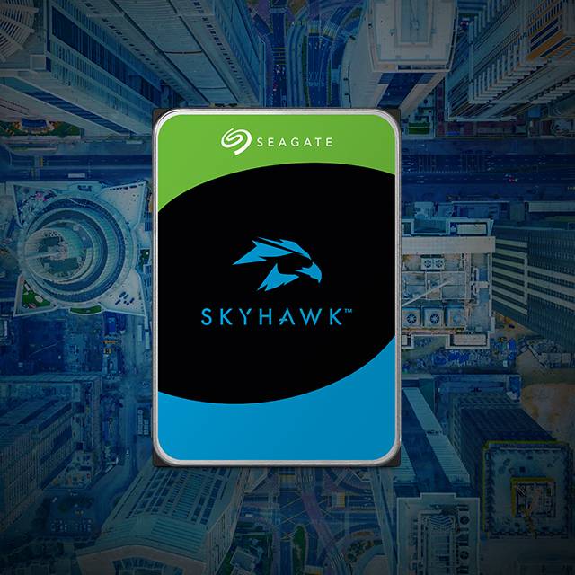 La imagen muestra un disco duro de la marca Seagate, modelo SkyHawk, con un diseño de fondo que parece una vista aérea de una ciudad. El disco duro tiene una etiqueta con los colores verde, negro y azul, y el logo de un halcón en el centro. La palabra SEAGATE está en la parte superior de la etiqueta, y SKYHAWK está en la parte inferior.