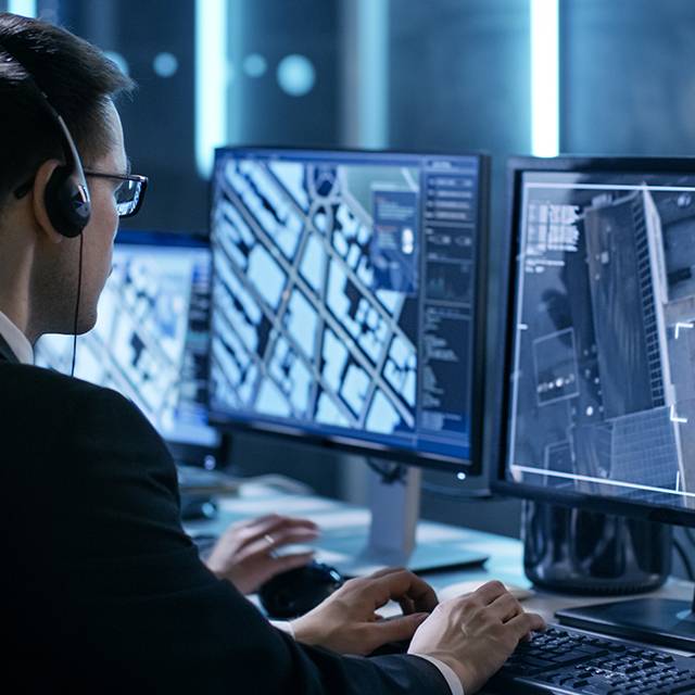 Persona trabajando en una computadora con múltiples monitores que muestran imágenes de mapas y datos de vigilancia.