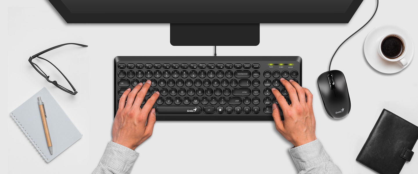 La imagen muestra un escritorio visto desde arriba. En él, hay un Teclado Genius SlimStar Q200 USB Negro con manos tecleando, un monitor de computadora frente al teclado, una taza de café a la derecha, gafas a la izquierda y un cuaderno con un bolígrafo encima, justo al lado de las gafas.