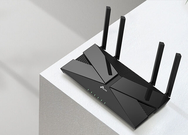Un router TP-Link Archer AX23 negro con cuatro antenas verticales sobre una superficie blanca. El router tiene un diseño angular con detalles brillantes y luces LED en el frente. A la derecha se ve parcialmente un libro o un objeto blanco.