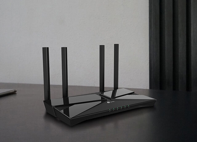 Un router TP-Link Archer AX23 negro con cuatro antenas verticales sobre una superficie negra brillante. A la izquierda se ve parcialmente un teléfono inteligente y un objeto blanco. El fondo muestra una pared gris con paneles verticales negros.